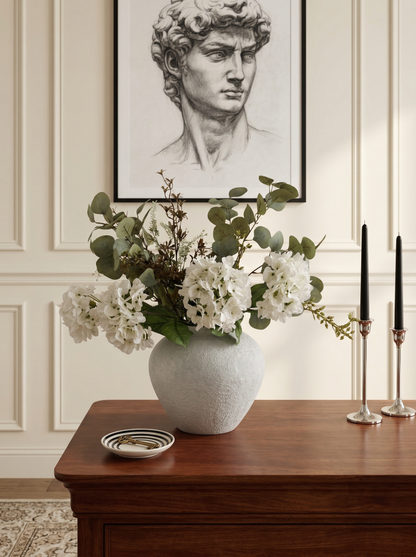 Decorative vase with flowers on a wooden table against a wall with a framed portrait.