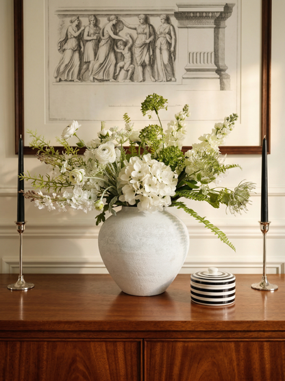 Floral arrangement in a white vase on a wooden surface with a classical painting in the background.