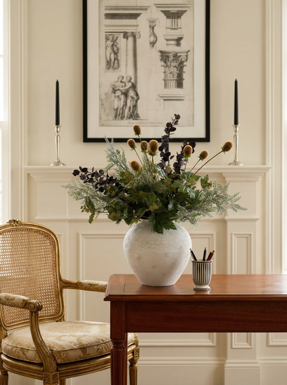 Floral arrangement in a white vase on a wooden table with a chair and framed artwork in the background.