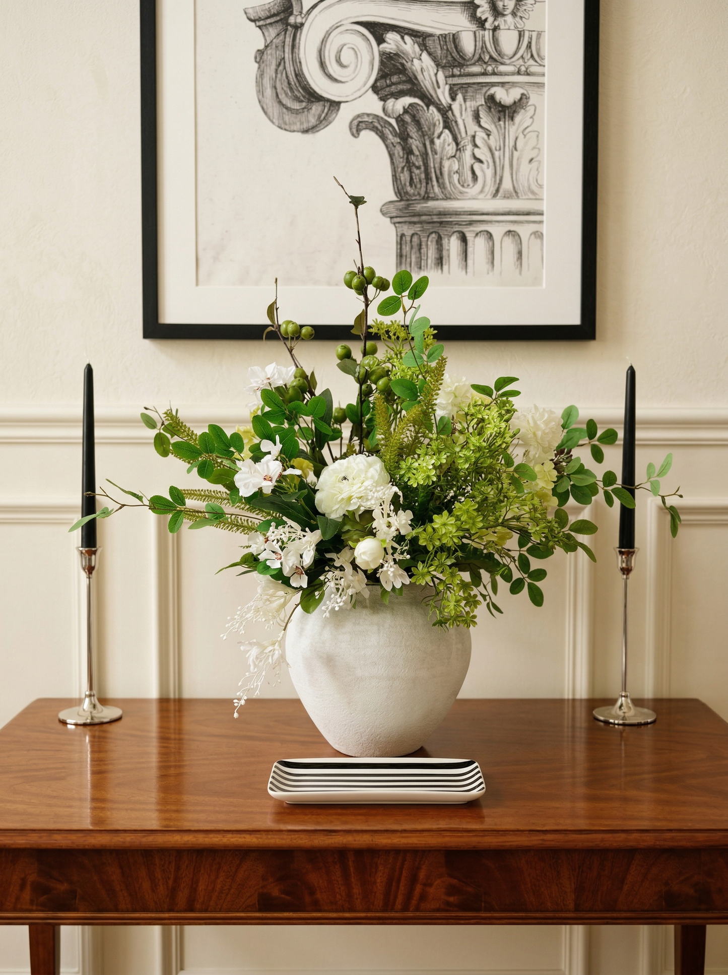 Floral arrangement in a white vase on a wooden table with black candles and a framed picture in the background.