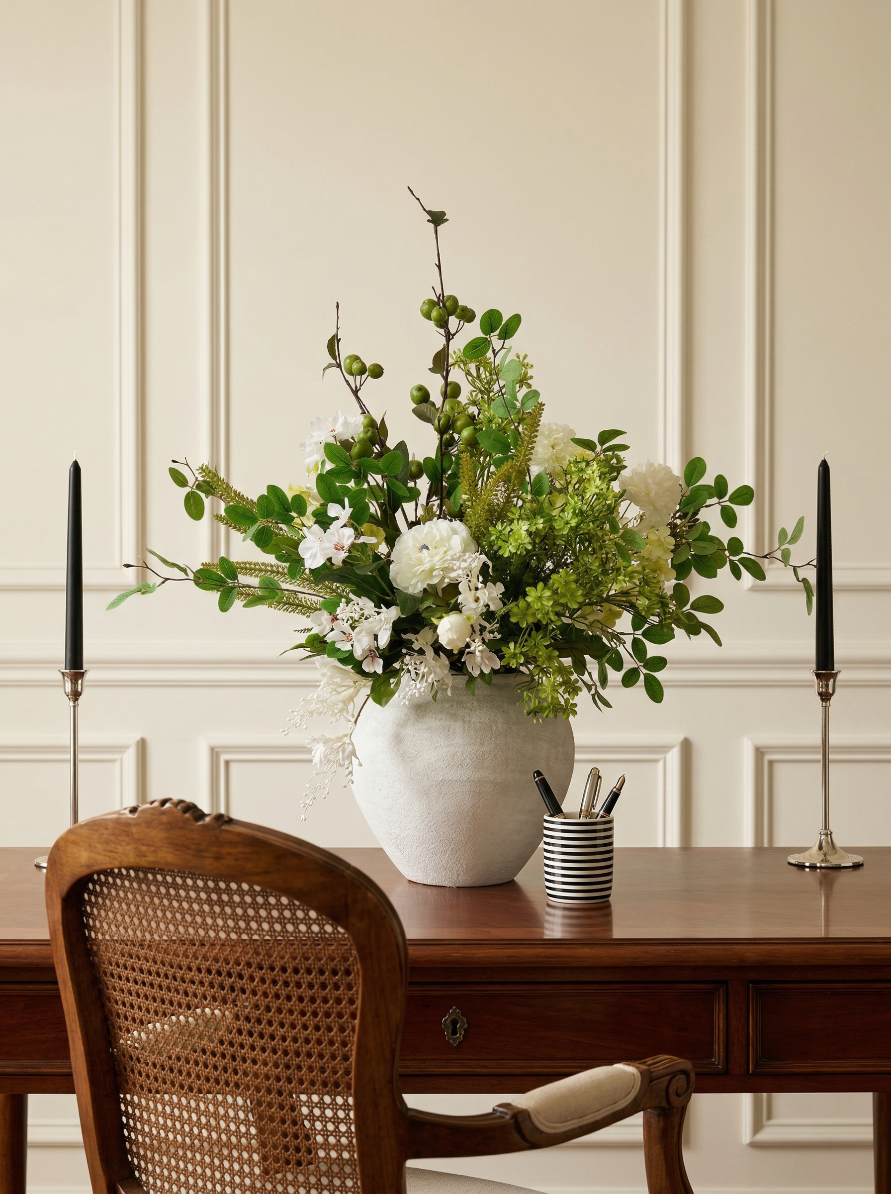 Floral arrangement in a white vase on a wooden table with candles and a chair.