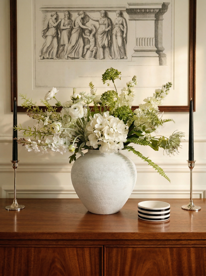 White vase with flowers on a wooden surface in front of a decorative wall piece.