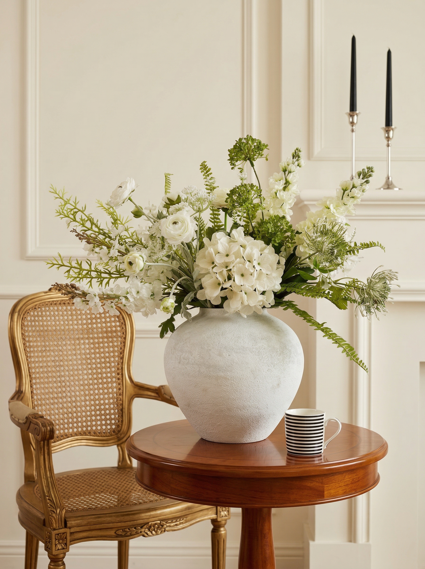 White vase with flowers on a wooden table next to a wicker chair in a room.