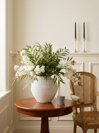 White vase with greenery on a wooden table in a room with a chair and candles.